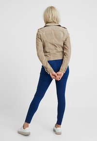Vrouw met blond haar, een beige jas, blauwe jeans en witte sneakers, staand met de handen achter de rug geklemd tegen een witte achtergrond.