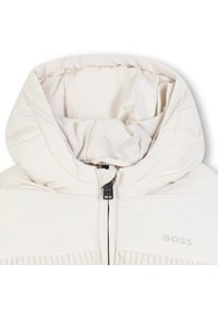 Giacca piumino beige chiaro con colletto alto, zip frontale e dettagli a costine. Presenta una finitura liscia e lucida e un dettaglio del logo.