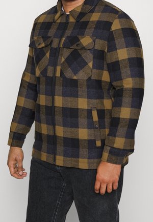 Mann trägt eine braun-schwarz karierte Reißverschlussjacke mit Klapptaschen auf der Brust und seitlichen Taschen mit Druckknopfverschluss, kombiniert mit dunklen Jeans.