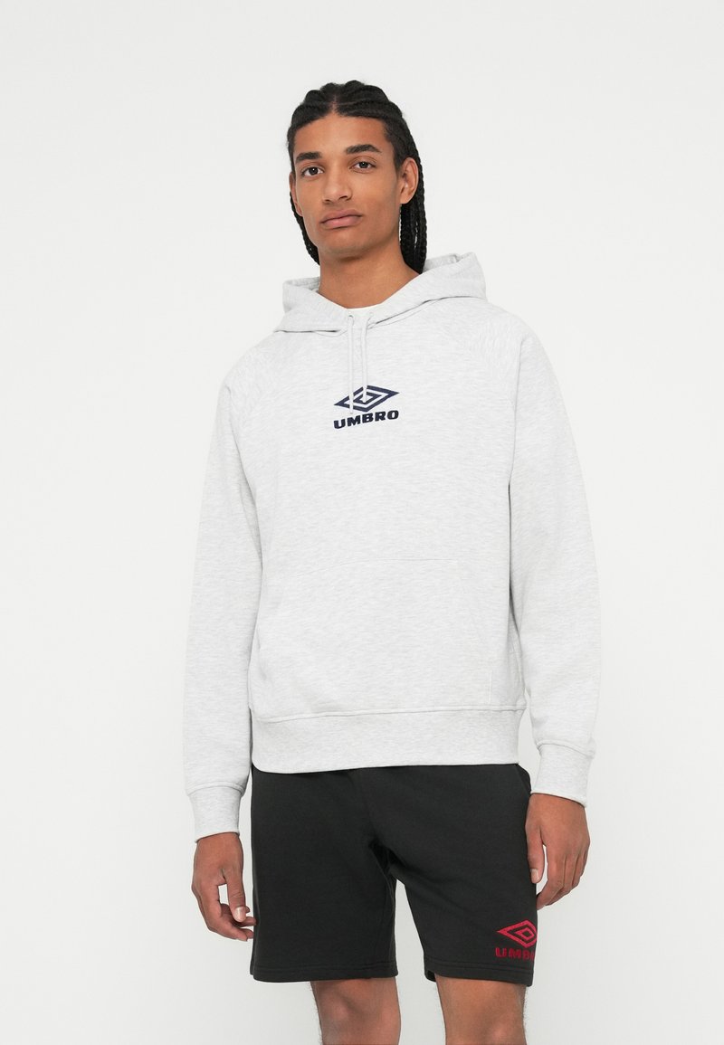 Joven con cabello trenzado que lleva una sudadera ligera gris de Umbro y pantalones cortos negros de Umbro, de pie contra un fondo blanco liso.