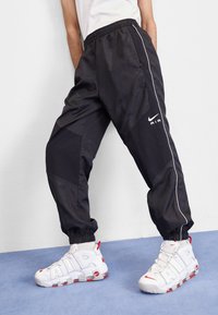 Schwarze Jogginghosen aus Nylon mit elastischem Bund und Bündchen am Knöchel, mit weißen Streifen und dem Nike Air Logo. Kombiniert mit weißen Sneakern.