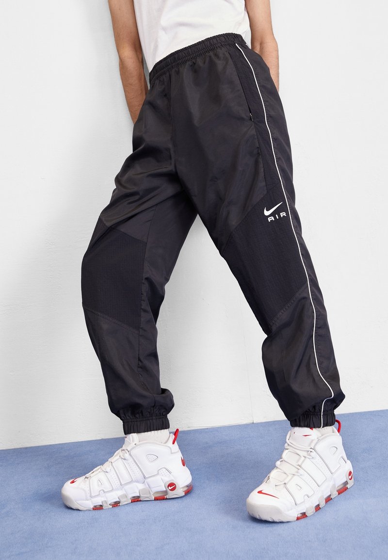 Schwarze Jogginghosen aus Nylon mit elastischem Bund und Bündchen am Knöchel, mit weißen Streifen und dem Nike Air Logo. Kombiniert mit weißen Sneakern.