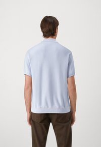 Kortärmad polo i ljusblått, gjord av texturerat tyg med ribbad krage och fåll, matchad med mörkbruna byxor.