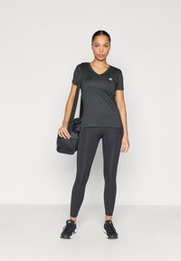 Czarna sportowa koszulka z dekoltem w serek i logo, w połączeniu z czarnymi legginsami i czarnymi trampkami, trzymająca czarną torbę sportową.