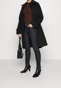 Kvinna i svart kappa, brun ribbad tröja, mörka skinny jeans, svarta ankelskor, håller en svart handväska med vävt mönster.