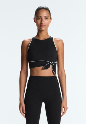 Kobieta ubrana w czarny, bezrękawnik crop top z białym obszyciem i wiązaniem po bokach, w połączeniu z czarnymi legginsami z wysokim stanem, stojąca na jednolitym tle.