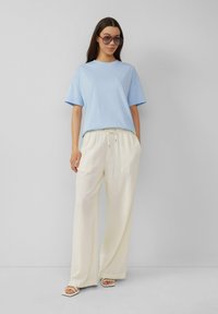 Lichtblauwe katoenen T-shirt met korte mouwen, gecombineerd met losse, crèmekleurige linnenbroeken. Model draagt grote, getinte zonnebril en witte sandalen.