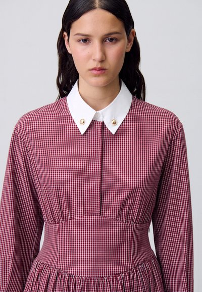 Modella con lunghi capelli scuri che indossa un vestito a quadretti rossi e bianchi con un colletto bianco, posata su uno sfondo neutro.
