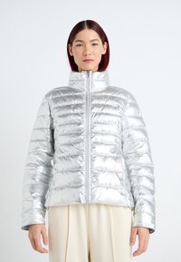 Giacca a vento argentata con colletto alto, caratterizzata da un design trapuntato, una texture lucida e chiusura con zip. Indossata sopra pantaloni beige.