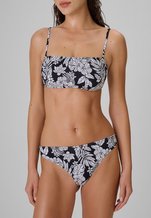Kobieta ubrana w czarny bikini z białym kwiatowym wzorem, cienkimi ramiączkami i majtkami o średnim stanie na jednolitym szarym tle.