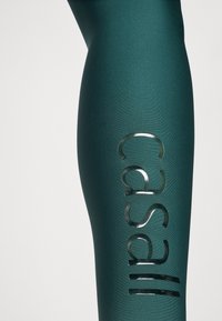 Leggings verdes de talle alto hechos de material elástico, con un logotipo plateado brillante de "Casall" en el muslo izquierdo. Textura suave.