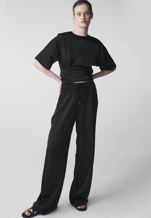 Młoda kobieta stojąca z rękami na biodrach, ubrana w luźną czarną koszulkę, szerokie czarne spodnie i czarne sandały na jednolitym tle.