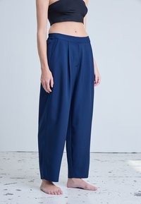 Femme portant un pantalon bleu marine ample et un crop top noir, debout pieds nus sur un sol blanc usé, devant un fond uni.