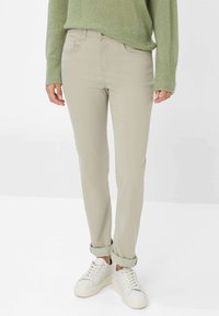 Persona che indossa pantaloni beige slim-fit arrotolati ai polsini, sneaker bianche e un maglione di lana verde, in piedi contro uno sfondo bianco.