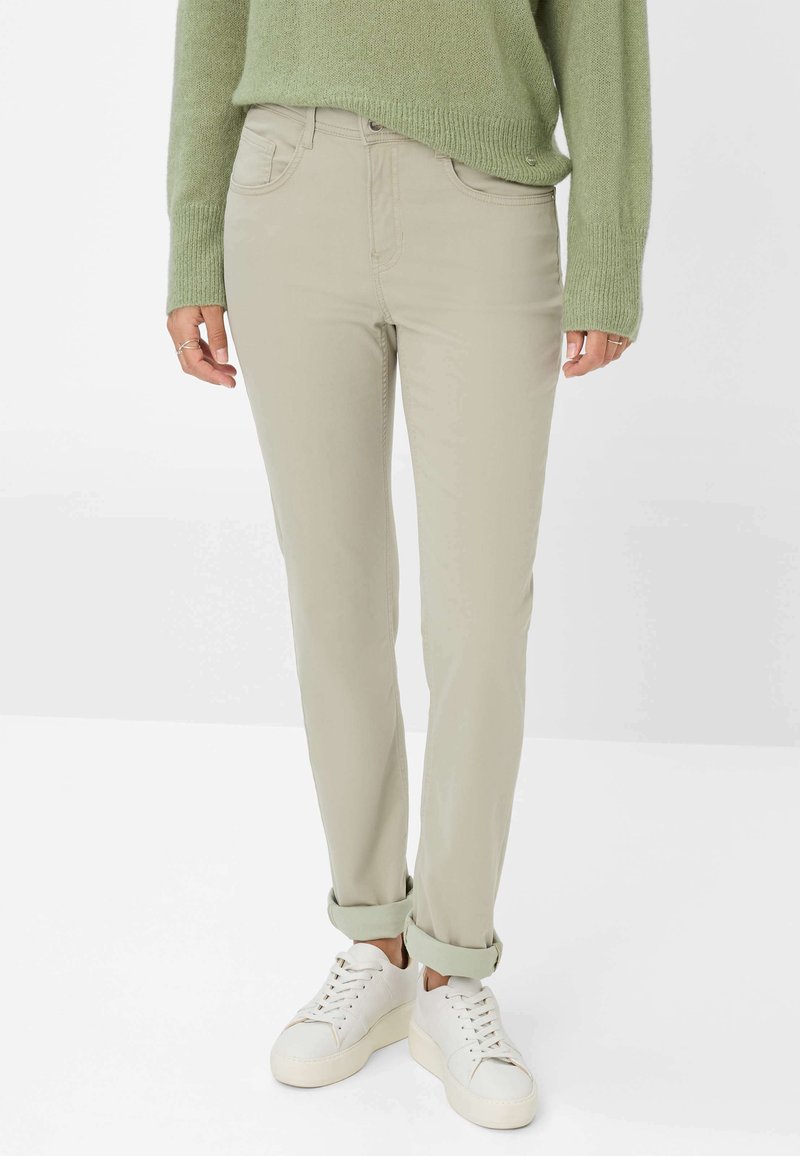 Persona che indossa pantaloni beige slim-fit arrotolati ai polsini, sneaker bianche e un maglione di lana verde, in piedi contro uno sfondo bianco.