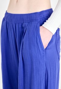 Pantalon léger bleu avec un tissu texturé, une ceinture élastique et des poches latérales. Le design présente une coupe décontractée et des coutures verticales.