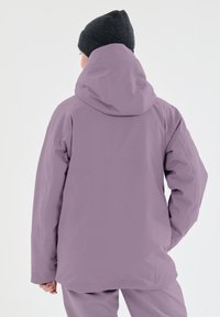Giacca impermeabile di un leggero viola con cappuccio, caratterizzata da una texture liscia, un design leggermente svasato e una vestibilità rilassata. Berretto in maglia nera visibile.
