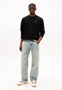 Sudadera negra, vaqueros anchos de pierna en azul claro, zapatillas blancas con detalles en negro. Estilo casual, marca mínima en la sudadera.