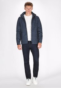 Marine gestreepte jas met hoge kraag, ritssluiting, gedragen over een wit shirt. Gecombineerd met donkere jeans en witte sneakers.