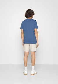 Blå bomull t-shirt med kort ärm, bärs med beige linnebyxor, stående figur vänd bort, i kombination med vita sneakers och ankelsockor.