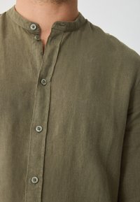 Olive green button-up shirt made of textured fabric. Features a band collar and two matching buttons at the neck. Simple design.