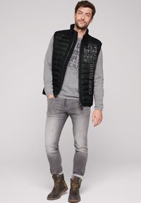 Gilet isolante nero sopra una felpa grigia con stampa grafica, abbinato a jeans grigi chiari e stivali marroni stringati.