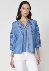 Ljust blå denimblus med trekvartsärmar, prydd med intrikata blå broderier, V-ringning och snörningsdetaljer.