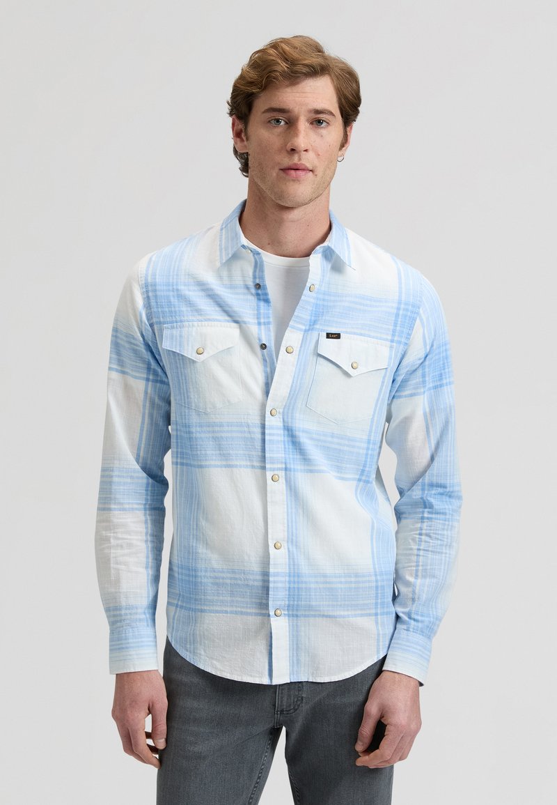 Νεαρός άνδρας που φοράει πουκάμισο με ανοιχτό μπλε και λευκό καρό πάνω από λευκό t-shirt και σκούρα γκρι τζιν, στέκεται μπροστά σε απλό φόντο.