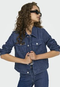 Giacca di jeans blu scuro con maniche lunghe, due tasche sul petto, bottoni argentati e colletto. Le pieghe aggiungono texture al tessuto.