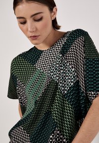 Lässiges Kurzarmoberteil mit geometrischem Muster in Grün, Schwarz und Weiß, das unterschiedliche Texturen und Formen im gesamten Stoff aufweist.