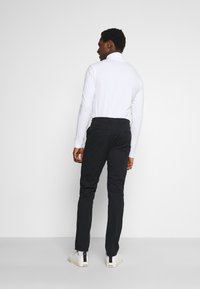 Pantalones ajustados negros con bolsillos traseros, combinados con una camisa blanca de manga larga. La persona lleva zapatillas blancas. Vista desde atrás.