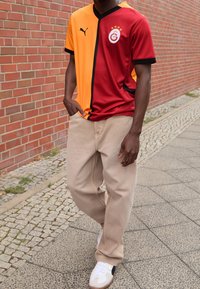 Camiseta de Puma con mitades en naranja y rojo, acentos en negro y logotipo del equipo. Combinada con pantalones beige y zapatillas blancas en una calle de adoquines.