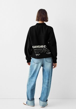 Sweatshirt noir oversize avec impression Kansas City au dos, associé à un jean baggy bleu clair. Tissu lisse et coupe décontractée.