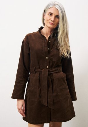 Robe en velours côtelé marron à manches longues, avec un devant à boutons et une taille cintrée par un lien. Elle possède deux poches avant et un design sans col.
