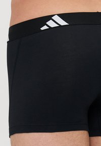 Svarta bomullskalsonger med en bred elastisk midja som har tre vita ränder på framsidan. Slät textur och åtsittande design.