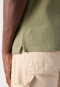 Un bras à la peau foncée à côté d'une chemise texturée couleur olive et d'un pantalon cargo beige avec une grande poche et des détails brodés.
