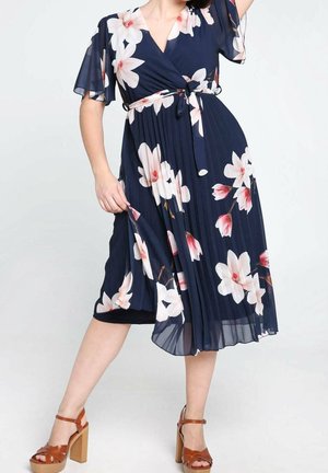 Frau trägt ein marineblaues plissiertes Midikleid mit weißem und pinkem Blumenmuster, braune Sandalen mit hohem Absatz, posiert mit einer Hand am Rock.