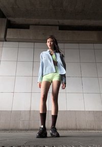 Ljusblå, texturerad skjorta med knappar över en limegrön bikini och shorts. Mörkbruna strumpor och rejält svarta sneakers fulländar outfiten.