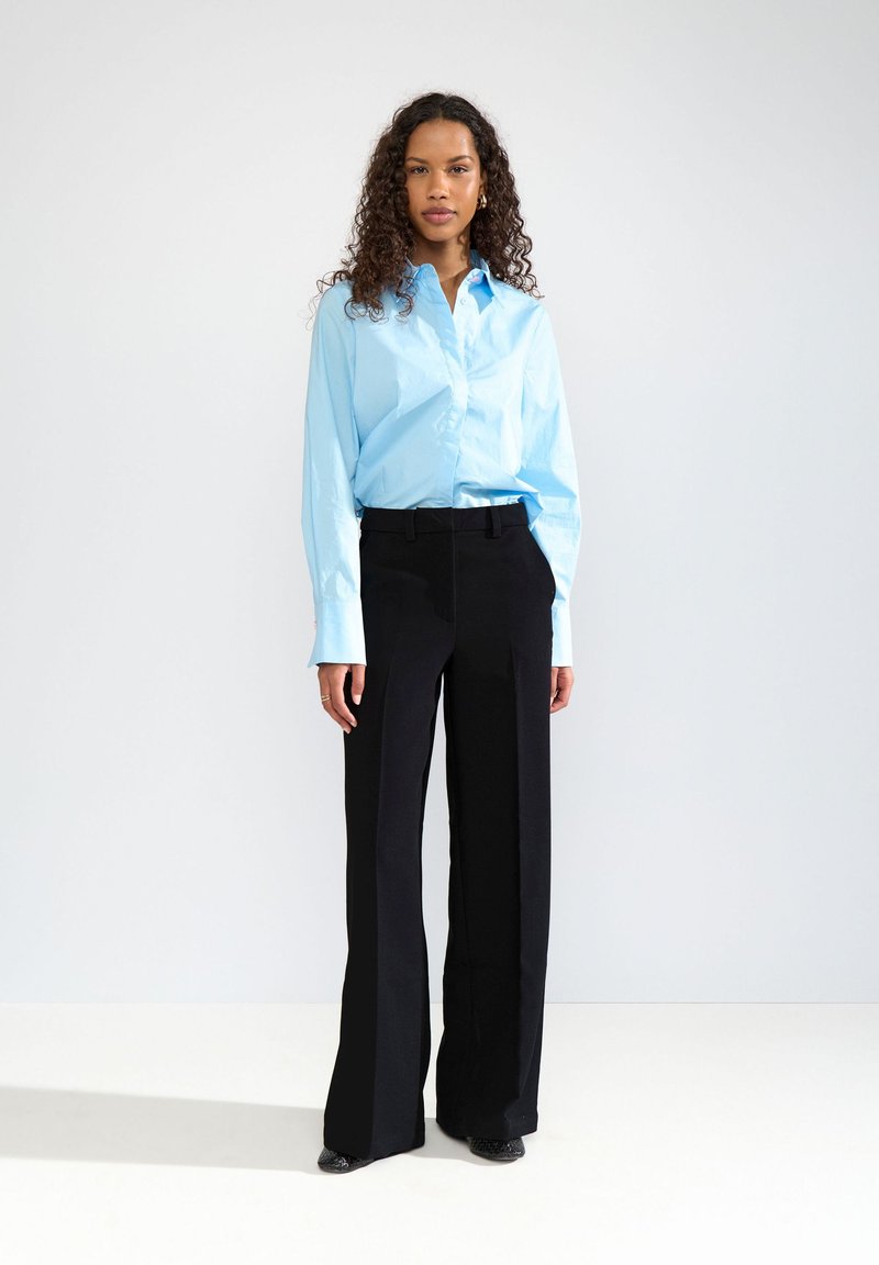 Femme aux cheveux bouclés portant une chemise bleu clair boutonnée et un pantalon large noir, debout devant un fond blanc.