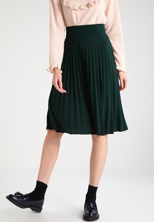 Femme portant une jupe plissée vert foncé, un chemisier rose clair à manches longues avec des volants, des chaussettes noires et des mocassins noirs à pompons.