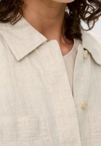 Zbliżenie na osobę w beżowej lnianej koszuli z kołnierzykiem i guzikami, pokazujące część ich szyi i kręcone włosy.