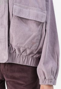Gros plan d'une personne portant une veste en velours côtelé violet clair avec une grande poche et des poignets élastiques, associée à un pantalon en velours côtelé violet foncé.