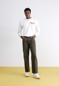 Chemise blanche à manches longues avec imprimé rouge, pantalon vert olive, et baskets blanches avec semelle en gomme. Le modèle se tient sur un sol jaune devant un fond uni.
