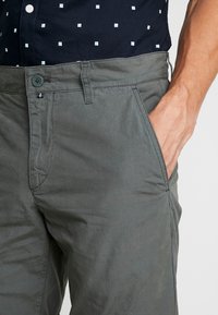La mano di un uomo nella tasca di pantaloni grigi indossati con una camicia blu navy con piccoli quadrati bianchi, mostrata dalla vita a metà coscia su sfondo bianco.