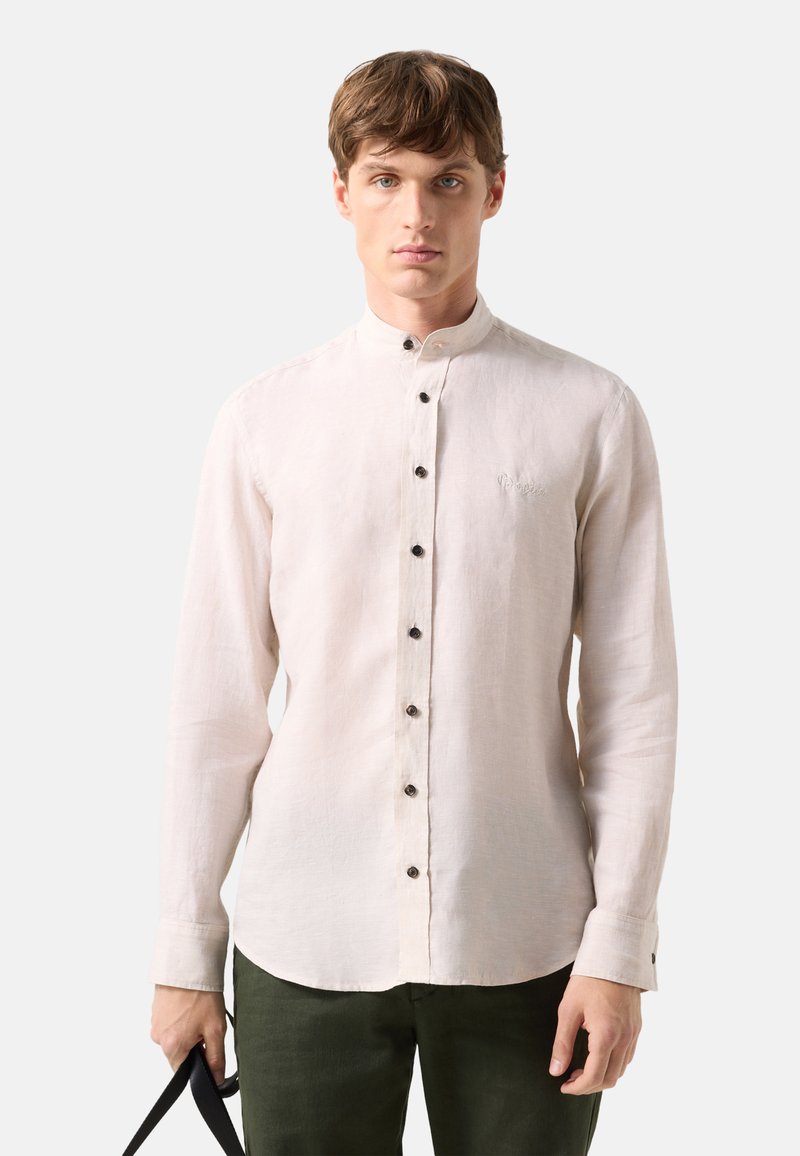Young man wearing a light beige Mandarin collar button-up shirt and dark green pants, holding a black strap in his left hand.