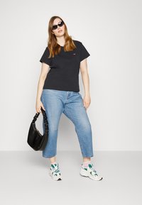 Fekete rövidujjú póló, világoskék cropped farmernadrág, fehér és zöld sportcipő, valamint egy fekete textil kézitáska. A napszemüveg teszi teljessé az öltözetet.