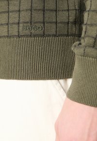 Gros plan sur le poignet et la taille d'un pull en maille vert olive à motif à carreaux et logo « HUGO », sur un pantalon blanc avec le poignet visible.