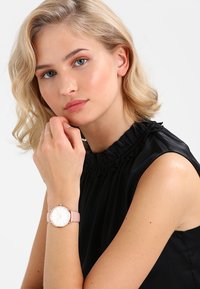 Orologio rotondo con cinturino in silicone rosa e hardware argentato, con quadrante bianco e indicatori delle ore minimalisti. Modella che indossa una maglia nera.