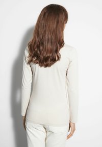 Długi rękaw, kremowy top z miękkiego materiału, dopasowany fason, gładka tekstura, z minimalnymi detalami szwów, noszony z jasnymi spodniami.