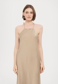Beige halter-neck dress in soft satin fabric, featuring a minimalist design and a flowing silhouette, complemented by gold-tone earrings.